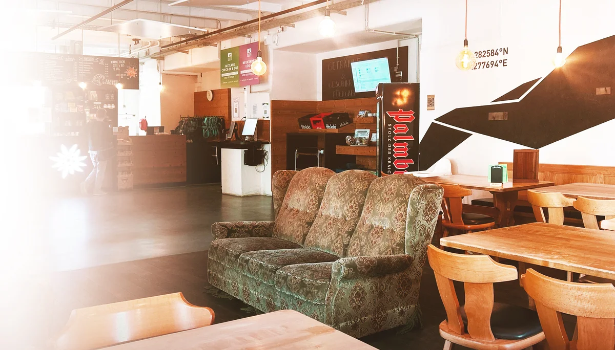 Überblick über das Bistro im Eingangsbereich: im Vordergrund mehrere Tische und Stühle aus Holz, ein grünes Sofa in der Mitte. im Hintergrund ein Kühlschrank und die Theke | © diekletterarena Bistro Gipfeltreff in der Kletterarena | © diekletterarena