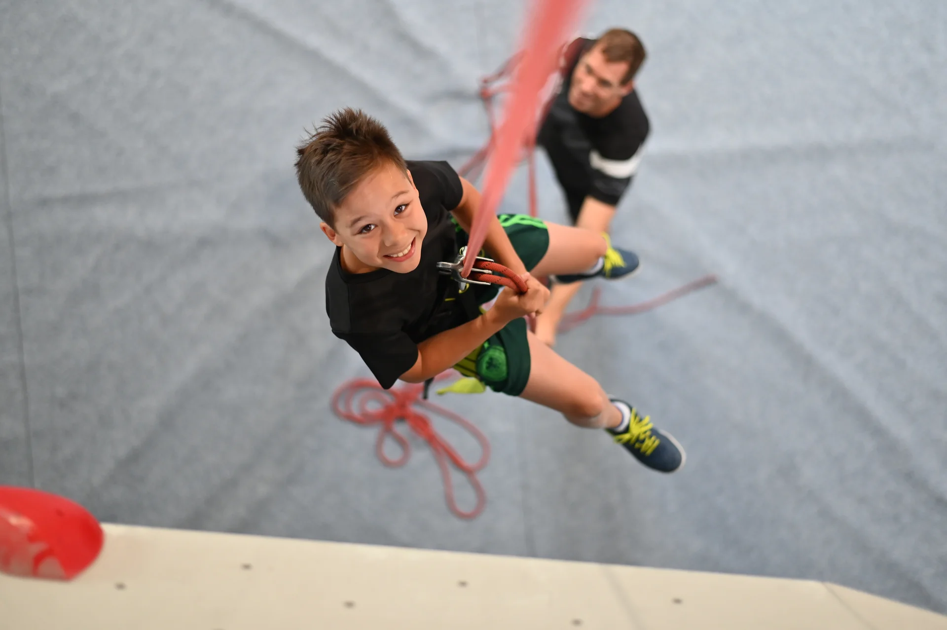 Kind hängt am Seil an der Kletterwand und lächelt | © diekletterarena Kind hängt am Seil an der Kletterwand und lächelt | © diekletterarena